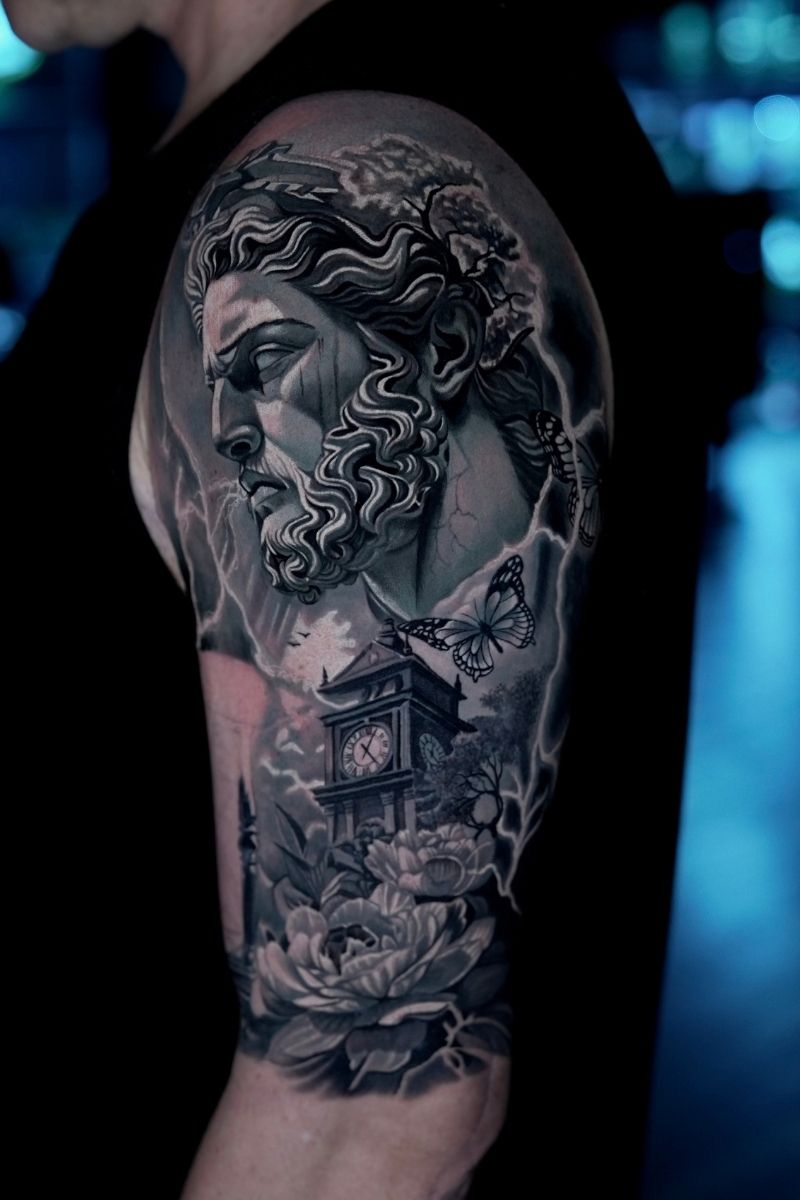 Black and grey upper arm tattoo featuring a sculptural face, butterflies, a clock tower, and flowers