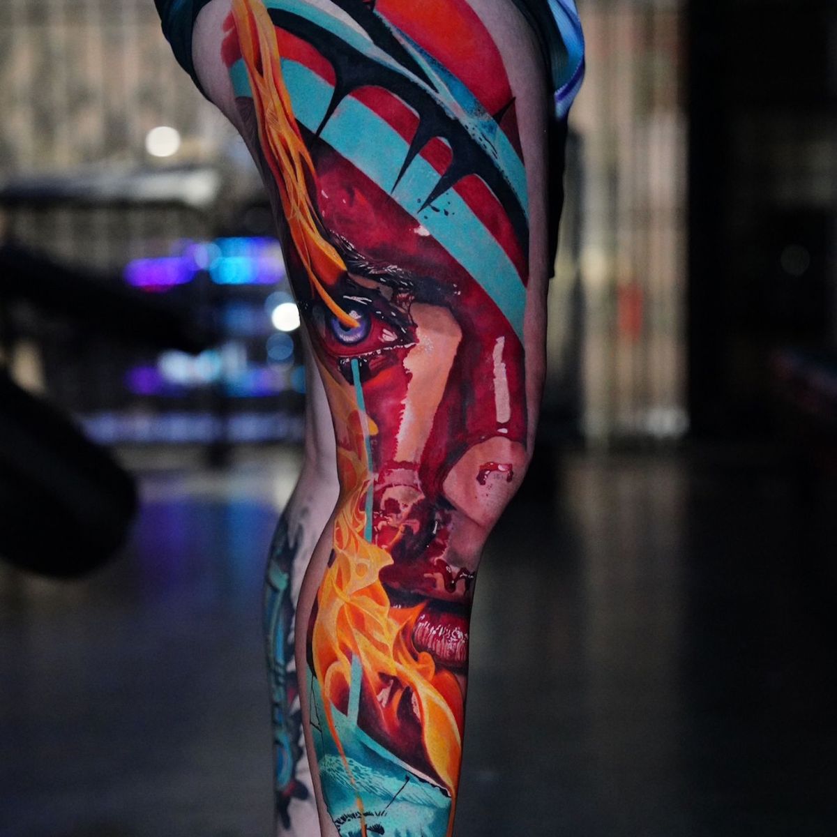 Color leg tattoo featuring a large face with vivid red, turquoise and flowing orange details by Frank Tattoo Arts